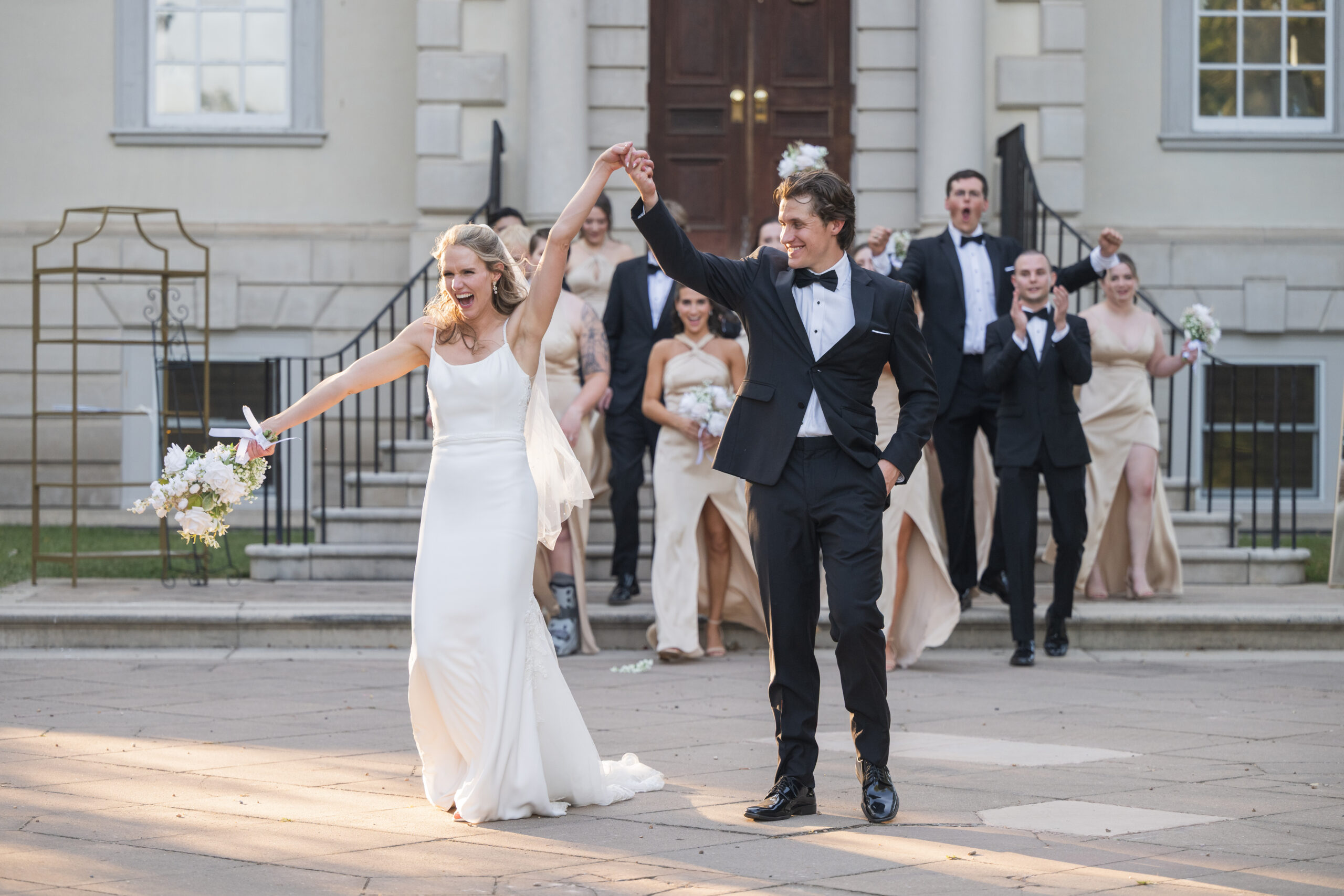 Joyful bride and groom holding hands and walking down the Georgian manor courtyard steps at Great Marsh Estate, followed by a cheering bridal party, photographed by Amberlee Christey Photography.